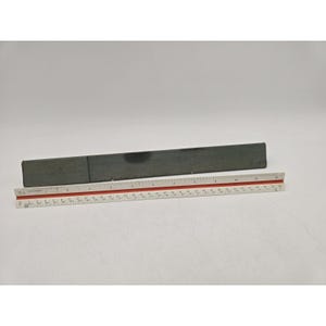 May include: A rectangular, dark gray metal bar with a smooth, matte finish positioned behind a white and red ruler. The ruler shows the metal bar measures approximately 12 inches in length. The bar has a visible seam and a dark circular mark.