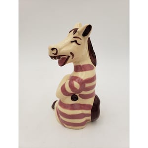 May include: A ceramic figurine of a smiling zebra with a cream-colored body and dark brown and maroon stripes. The zebra is in a seated position with its arms crossed, and its tongue is sticking out. The figurine has a glossy finish.