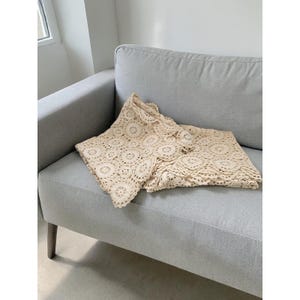 May include: A cream-colored, crocheted blanket and matching shorts set. The blanket features a circular pattern with a scalloped edge. The shorts are folded neatly next to the blanket on a gray sofa. The set is made of a soft, textured material.