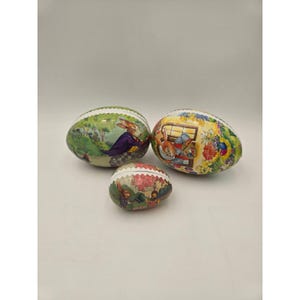 May include: Three decorative Easter egg-shaped tins. The tins feature colorful illustrations of rabbits in various scenes, including a rabbit in a purple coat, and rabbits near a window. The tins are green, white, and red.