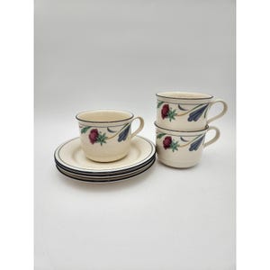 May include: A set of cream-coloured teacups and saucers with a floral pattern. The cups and saucers have a black trim. The floral design features red flowers and blue leaves. Two cups are stacked, and the other cup and saucers are separate.