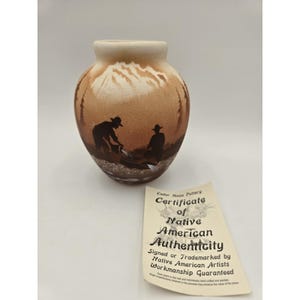May include: A tan and brown ceramic vase featuring a painted silhouette of two figures panning for gold in a mountain landscape. Beside the vase is a paper Certificate of Native American Authenticity from Cedar Mesa Pottery with black text.