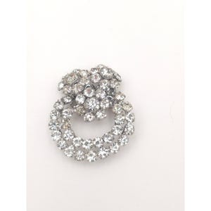 May include: A silver-toned brooch with a double-loop design, encrusted with numerous clear, sparkling rhinestones. The brooch has a cluster of rhinestones at the top, adding a touch of elegance to the jewelry piece.