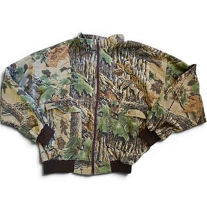 May include: A zip-up jacket featuring a realistic forest camouflage pattern with brown tree bark, green leaves, and branches. The jacket includes a high collar, a dark brown front zipper, two large front pockets with flaps, and dark brown ribbed cuffs.