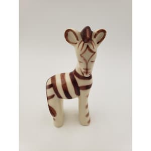 May include: A ceramic zebra figurine with a cream-colored body and dark brown stripes. The zebra has a long neck, small ears, and a stylized face. The figurine is standing upright and appears to be handmade.