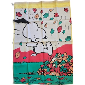 May include: A rectangular fabric wall hanging featuring a cartoon illustration of Snoopy jumping into a large pile of colorful autumn leaves. The background includes a yellow top border, a red middle section, and a green bottom section.