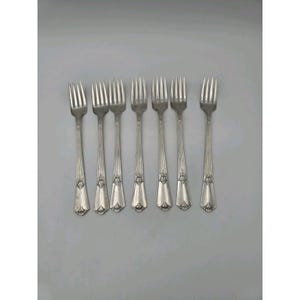May include: A set of seven silver-toned metal forks arranged in a row. Each fork features a slender handle with vertical fluting and a decorative embossed floral motif near the base of the handle, leading to a four-tined head.