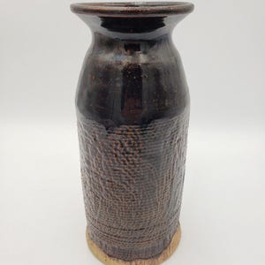 May include: A dark brown ceramic vase with a textured surface and a slightly flared opening. The vase has a light brown rim and base.
