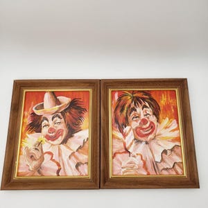May include: Two framed paintings of clowns. Each painting depicts a clown with red hair, a white ruffled collar, and a red nose. One clown wears a hat and holds a flower, the other holds a feather. The frames are brown with gold inner edges.