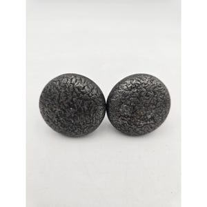 May include: Two round, textured, dark gray cabinet knobs. The knobs have a rough, pitted surface, and are set against a white background. These decorative knobs are suitable for furniture or cabinetry.