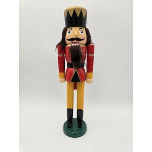 May include: A wooden nutcracker figurine, painted in red, black, gold, and yellow. The figurine has a black hat with gold accents, a red jacket, and yellow legs. It stands on a green base.