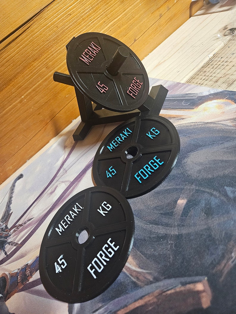 Op de afbeelding: Drie zwarte gewichtsplaten met de woorden "MERAKI" en "FORGE" erop gedrukt. De platen zijn elk 45 kg.