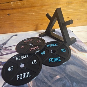 Op de afbeelding: Vier zwarte gewichtsplaten met de tekst "MERAKI FORGE 45 KG" erop gedrukt. De platen zijn op elkaar gestapeld en op de achtergrond staat een zwarte metalen standaard.
