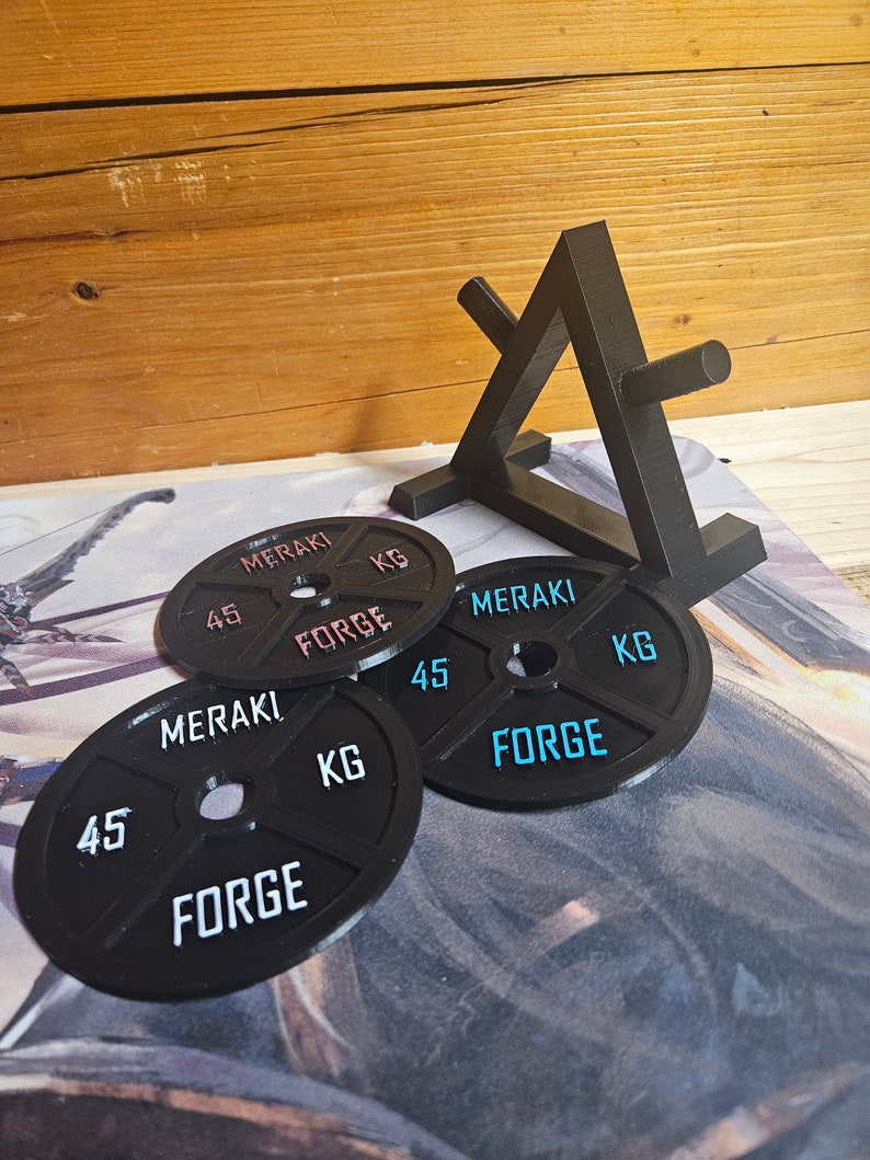 Op de afbeelding: Vier zwarte gewichtsplaten met de tekst "MERAKI FORGE 45 KG" erop gedrukt. De platen zijn op elkaar gestapeld en op de achtergrond staat een zwarte metalen standaard.