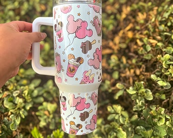 May include: White stainless steel travel mug with a pink and white Mickey Mouse themed design. The mug has a handle and a lid. The design features Mickey Mouse ears, balloons, popcorn, ice cream, and other Disney-themed treats.