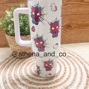 Op de afbeelding: Een witte roestvrijstalen beker met een wit deksel en handvat. De beker heeft een herhalend patroon van rode en blauwe cartoon Spider-Man-personages met een wit spinnenweb en roze strikken. De beker heeft ook een roze Hello Kitty-personage met een wit hart.