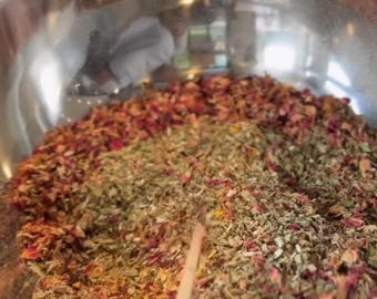 May include: A stainless steel bowl filled with a mixture of dried herbs and flower petals. The mixture is primarily composed of green, brown, and red elements, with a wooden stirring stick visible. The contents appear to be a blend of botanicals.