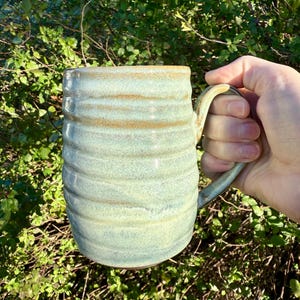 May include: A handmade ceramic mug with a ribbed design. The mug has a light blue and tan glaze, and a large handle. The mug is held in a hand against a background of green foliage. This mug is perfect for coffee, tea, or any hot beverage.