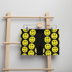 May include: Black boxer briefs with a repeating pattern of yellow smiley faces with dollar sign eyes. The underwear is hanging on a wooden hanger against a white background.