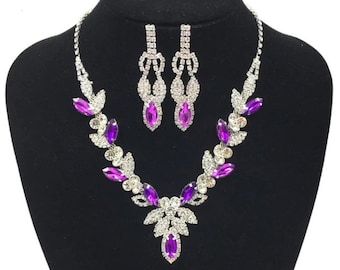 May include: A silver necklace and earrings set with purple and clear rhinestones. The necklace features a leaf design with a pendant in the center. The earrings are dangle style with a leaf design.