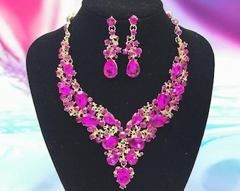 May include: A pink rhinestone necklace and earrings set. The necklace is a v-shaped design with a teardrop pendant. The earrings are teardrop shaped.