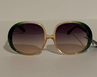 May include: Oversized sunglasses with a gradient lens. The frames are a light tan color with green accents on the top of the frames. The lenses transition from a dark purple to a light brown.
