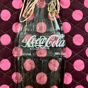 May include: A clear glass Coca-Cola bottle ornament with a straw-like bow on the top. The bottle is sitting on a brown and pink polka dot fabric.