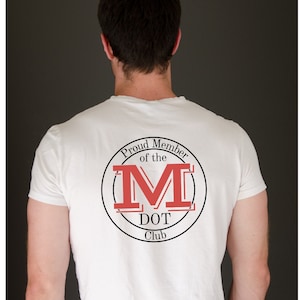 May include: A white short-sleeved t-shirt featuring a circular graphic on the back. The design includes the text "Proud Member of the M DOT Club" in black lettering, surrounding a large, bold, red letter M with black outlines and horizontal lines.