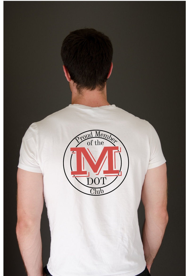 May include: A white short-sleeved t-shirt featuring a circular graphic on the back. The design includes the text "Proud Member of the M DOT Club" in black lettering, surrounding a large, bold, red letter M with black outlines and horizontal lines.