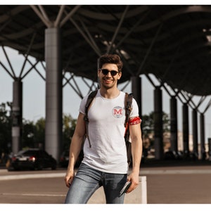 May include: A white short-sleeved t-shirt featuring a circular red and white logo with the letter M in the center, worn with blue jeans, a dark backpack, and a red patterned armband. The shirt includes small text around the central letter M.