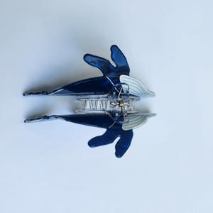 May include: A blue and white plastic hair clip with a clear plastic spring mechanism. The clip is shaped like a butterfly with a wavy design.