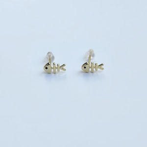 May include: A pair of gold-colored stud earrings in the shape of a fish skeleton. Each earring features a small, round head with a black dot for the eye and a series of horizontal lines representing the bones. The earrings are set against a white background.