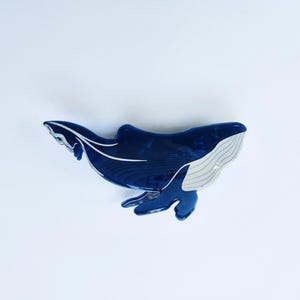 May include: A blue whale hair clip with a white underbelly. The whale is in a swimming position with its tail raised.