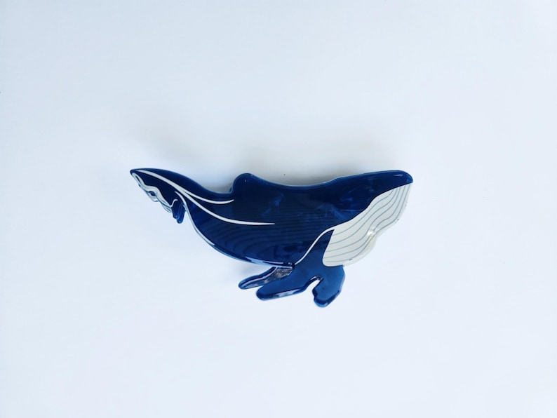 May include: A blue whale hair clip with a white underbelly. The whale is in a swimming position with its tail raised.