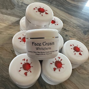 May include: A stack of white jars with red splatter designs. The top jar has a label that reads "Face Cream Whitefarm" and lists ingredients: "Water, Shea Butter, Hyaluronic Acid, Collagen, Dragon's Blood, Vitamin E".
