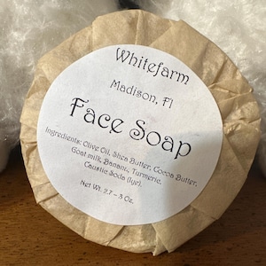 May include: Round face soap wrapped in brown paper. The label reads "Whitefarm, Madison, Fl, Face Soap." Ingredients include olive oil, shea butter, cocoa butter, goat milk, banana, turmeric, and caustic soda. Net weight is 2.7-3 oz.