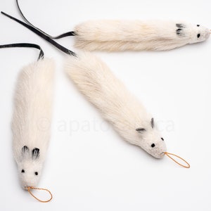 Puede incluir: Tres juguetes blancos para gatos con forma de ratón, cada uno con un cuerpo esponjoso, orejas negras y una cola negra. Los ratones tienen pequeños ojos negros y un lazo de cuerda naranja en la nariz.