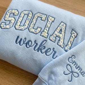 May include: Light blue sweatshirt with the words "SOCIAL worker" embroidered in dark blue. The word "SOCIAL" is in a floral pattern. The sleeve has the name "Emma" embroidered with a bow. A cozy, personalized gift.