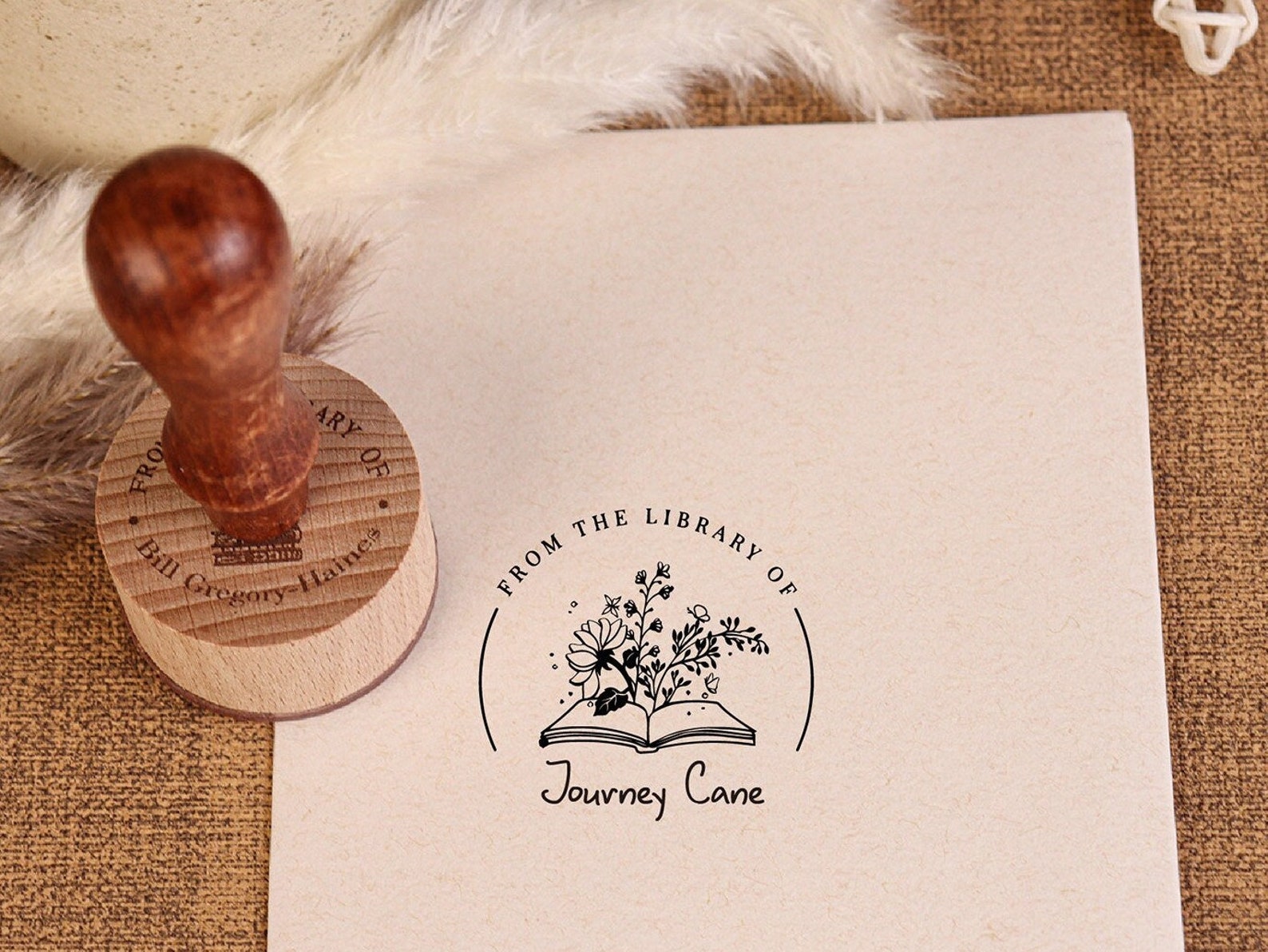 A piece of paper with a book stamp on it. The book stamp is an image of an open book with flowers coming out of it. A wooden book stamp and feather are next to the paper.