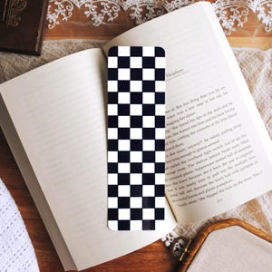 Può includere: Un segnalibro rettangolare caratterizzato da un motivo a scacchi bianco e nero ripetuto. Il segnalibro è posizionato verticalmente al centro di un libro aperto con pagine bianche e testo stampato in nero, mostrando un classico design a griglia.