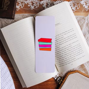 Könnte beinhalten: Rechteckiger Lesezeichen mit einer Abbildung eines Stapels bunter Bücher in Rot, Türkis, Pink und Orange. Das Lesezeichen liegt auf einem offenen Buch. Der Hintergrund umfasst eine Holzoberfläche und Spitze.