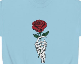 May include: Pale blue sweatshirt with a graphic of a skeleton hand holding a red rose. The rose has green leaves and a detailed design. The sweatshirt is a solid pale blue colour, with a crew neck and long sleeves.