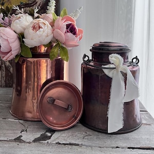 May include: Two copper containers with lids, one with a white ribbon, and a bouquet of pink flowers in the background.