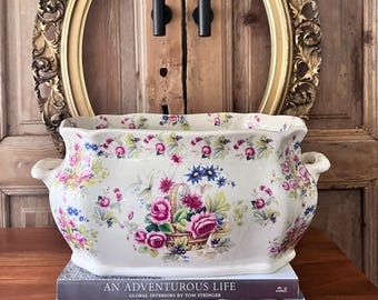 May include: A cream-colored, rectangular planter with a floral design featuring pink roses and a basket of flowers. The planter sits atop two books, one titled "An Adventurous Life." A gold-framed oval mirror hangs in the background.
