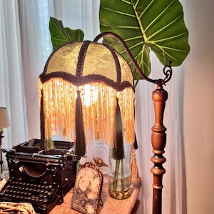 May include: A vintage-style floor lamp with a green lampshade trimmed with dark brown and gold fringe. The lamp has a wooden base and a curved arm. A large green leaf is behind the lamp. A vintage typewriter and a framed picture are on the table.