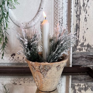May include: A lit white candle in a small, aged, beige-colored pot, surrounded by frosted evergreen branches. The pot has a textured, weathered appearance. The candle is centered, with the flame visible. The overall theme is rustic and decorative.