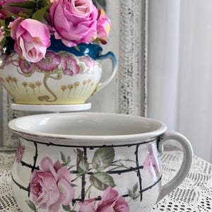 May include: A white ceramic planter with a handle, adorned with pink roses and green leaves, set against a black grid pattern. The planter is filled with pink roses. A second floral container is in the background.