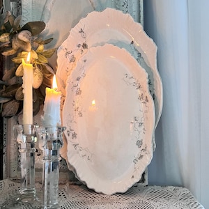 May include: Two white oval platters with a floral design and scalloped edges are stacked. Two lit candles sit in clear glass candle holders. The platters are on a lace tablecloth.