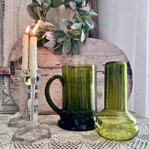 May include: A still life arrangement featuring a clear glass candlestick with lit candles, a green glass mug, and a green vase. A decorative wreath and lace tablecloth complete the scene.