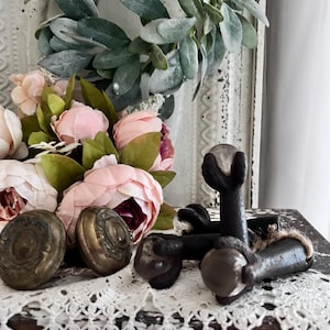 May include: An assortment of antique bronze and dark metal hardware, including door knobs and decorative feet. A bouquet of pink peonies and a green leaf wreath are in the background, all arranged on a white lace doily.